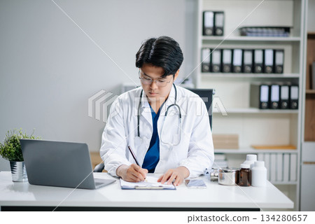 Confident young Asian male doctor in white medical uniform sit at desk working on computer. Smiling use laptop write 134280657
