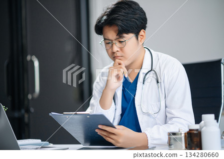 Confident young Asian male doctor in white medical uniform sit at desk working on computer. Smiling use laptop write Confident young Asian male doctor in white medical uniform sit at desk working on computer. Smiling use laptop write 134280660