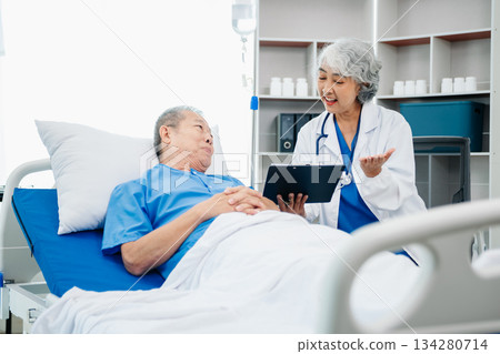 Asian doctor in white suit take notes while discussing and Asian elderly, man patient who lying on bed with receiving saline solution 134280714