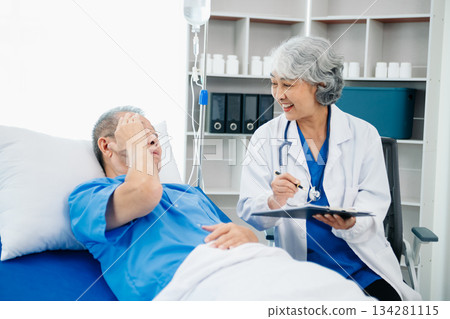 Asian doctor in white suit take notes while discussing and Asian elderly, man patient who lying on bed with receiving saline solution 134281115