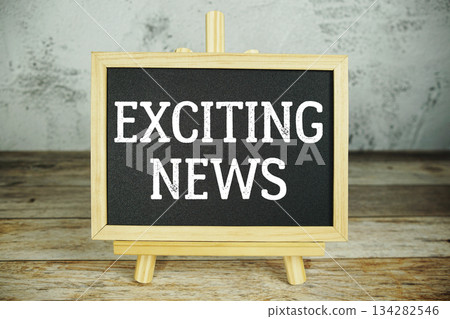 wooden easel displays a blackboard with the bold white text "Exciting News" set against a rustic wooden surface and textured gray wall background 134282546