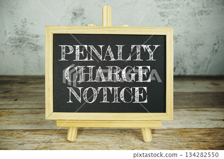 wooden easel displays a blackboard with the bold white text "Penalty Charge Notice" set against a rustic wooden surface and textured gray wall background 134282550