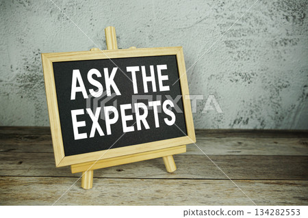 wooden easel displays a blackboard with the bold white text "Ask the Experts" set against a rustic wooden surface and textured gray wall background 134282553