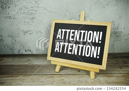 wooden easel displays a blackboard with the bold white text "Attention! Attention!" set against a rustic wooden surface and textured gray wall background 134282554