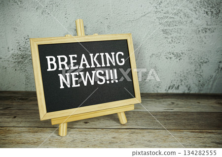wooden easel displays a blackboard with the bold white text "Breaking News" set against a rustic wooden surface and textured gray wall background 134282555