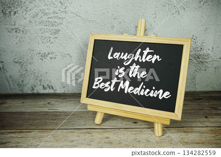 wooden easel displays a blackboard with the bold white text "Laughter is the Best Medicine" set against a rustic wooden surface and textured gray wall background 134282559