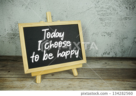 wooden easel displays a blackboard with the bold white text "Today I Choose to be Happy" set against a rustic wooden surface and textured gray wall background 134282560