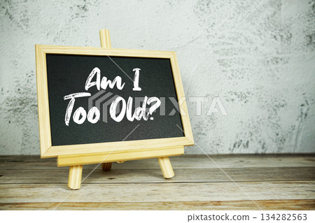 wooden easel displays a blackboard with the bold white text "Am I Too Old?" set against a rustic wooden surface and textured gray wall background 134282563