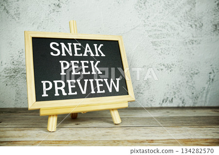 wooden easel displays a blackboard with the bold white text "Sneak Peek Preview" set against a rustic wooden surface and textured gray wall background 134282570