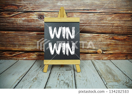 wooden easel holds a blackboard with the white text "Win-Win" set against a rustic wooden wall and light-colored wooden floor 134282583
