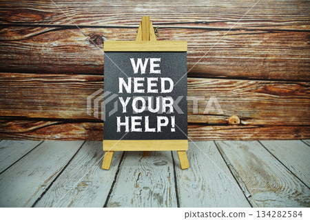 wooden easel holds a blackboard with the white text "We Need Your Help" set against a rustic wooden wall and light-colored wooden floor 134282584