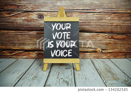 wooden easel holds a blackboard with the white text "Your Vote is Your Voice" set against a rustic wooden wall and light-colored wooden floor 134282585