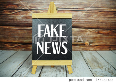 wooden easel holds a blackboard with the white text "Fake News" set against a rustic wooden wall and light-colored wooden floor 134282588