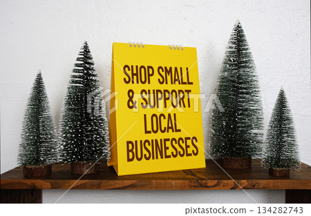 A bright yellow sign with the message "Shop Small & Support Local Businesses" sits on a wooden shelf, surrounded by miniature pine trees and a small decorative white house, creating a festive and 134282743