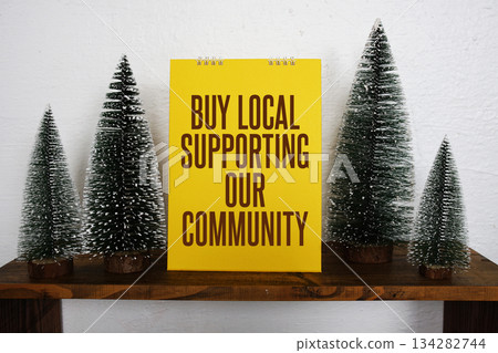 A bright yellow sign with the message "Buy Local Supporting Our Community" sits on a wooden shelf, surrounded by miniature pine trees and a small decorative white house, creating a festive and 134282744