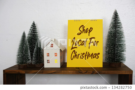 A bright yellow sign with the message "Shop Local For Christmas" sits on a wooden shelf, surrounded by miniature pine trees and a small decorative white house, creating a festive and supportive A bright yellow sign with the message "Shop Local For Christmas" sits on a wooden shelf, surrounded by miniature pine trees and a small decorative white house, creating a festive and supportive 134282750