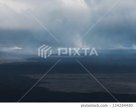 Dramatic aerial view of dark lava fields in Iceland, with steam rising from geothermal activity and heavy clouds creating a moody atmosphere. 134284480