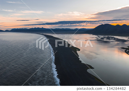 Aerial view of a black sand beach in Iceland, bordered by the ocean and a reflective water body, with distant mountains under a colorful sunset sky. Aerial view of a black sand beach in Iceland, bordered by the ocean and a reflective water body, with distant mountains under a colorful sunset sky. 134284482