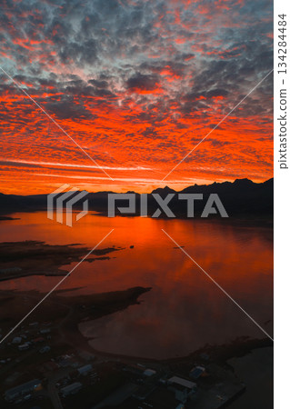 A vivid sunset with red and orange hues reflects on calm water, with silhouetted mountains and a small shoreline settlement in Iceland. A vivid sunset with red and orange hues reflects on calm water, with silhouetted mountains and a small shoreline settlement in Iceland. 134284484