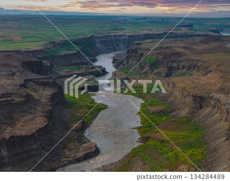 Studlagil Canyon in Iceland features steep basalt columns, a winding river, and green vegetation under a serene sunset sky. 134284489