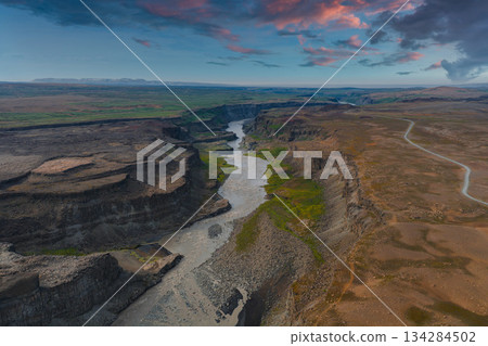 Studlagil Canyon in Iceland features basalt columns, a winding river, rocky terrain, green vegetation, and a vibrant pink and blue sky. Studlagil Canyon in Iceland features basalt columns, a winding river, rocky terrain, green vegetation, and a vibrant pink and blue sky. 134284502