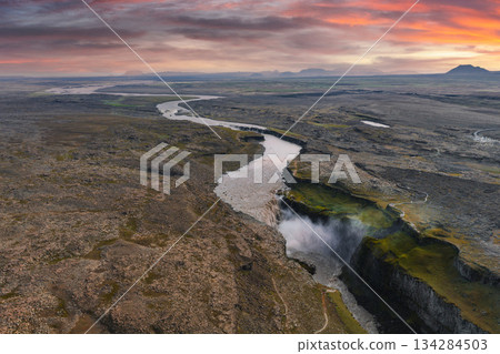 Aerial view of Dettifoss waterfall in Iceland, with mist rising from cascading water, a rugged canyon, a winding river, and a colorful sunrise sky. 134284503
