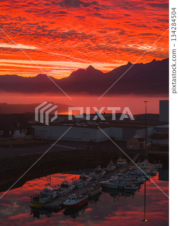 A vibrant sunset reflects on calm harbor waters in Iceland, with docked boats in the foreground and misty jagged mountains in the background. A vibrant sunset reflects on calm harbor waters in Iceland, with docked boats in the foreground and misty jagged mountains in the background. 134284504