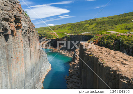 Studlagil Canyon in Iceland features geometric basalt columns, a turquoise river, green hills, and a dirt path under a blue sky with white clouds. 134284505