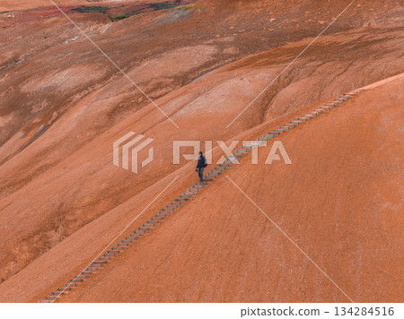 A lone figure walks up a staircase through a rust colored geothermal terrain in Iceland, with undulating surfaces and contrasting green patches in the background. 134284516