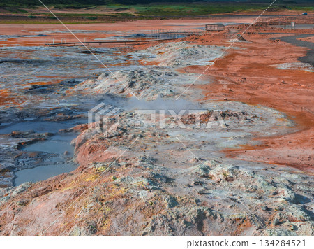 Rugged geothermal terrain in Iceland featuring red and orange soil, steaming vents, mud pots, mineral deposits, and a wooden walkway with green hills beyond. 134284521