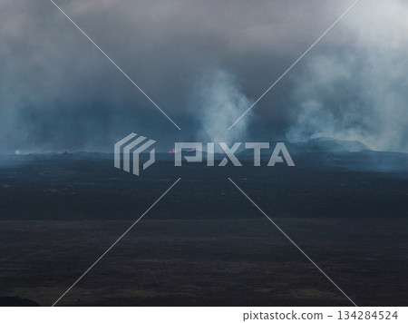 An active volcanic scene in Iceland shows smoke plumes, glowing lava, and rugged terrain under an overcast sky, highlighting geothermal activity. 134284524