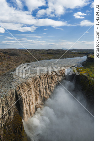Dettifoss waterfall in Iceland cascades over a rugged cliff into a mist filled canyon, surrounded by rocky terrain under a bright blue sky with clouds. 134284532