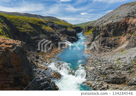 Studlagil Canyon in Iceland features a vibrant blue river, rugged basalt columns, and green vegetation under a clear blue sky, showcasing unique geology. Studlagil Canyon in Iceland features a vibrant blue river, rugged basalt columns, and green vegetation under a clear blue sky, showcasing unique geology. 134284548
