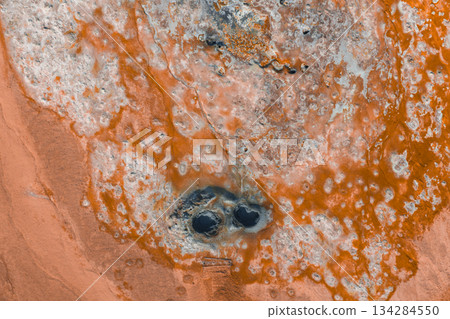 Aerial image of a geothermal area in Iceland with orange and white mineral deposits, textured surfaces, and two dark circular mud pots at the center. Aerial image of a geothermal area in Iceland with orange and white mineral deposits, textured surfaces, and two dark circular mud pots at the center. 134284550