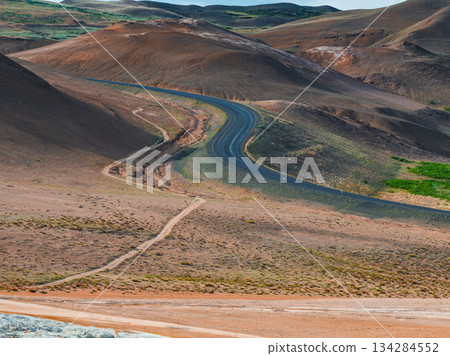 A winding road traverses a barren, reddish brown volcanic terrain in Iceland, with sparse vegetation, rolling hills, and a parallel dirt path. 134284552