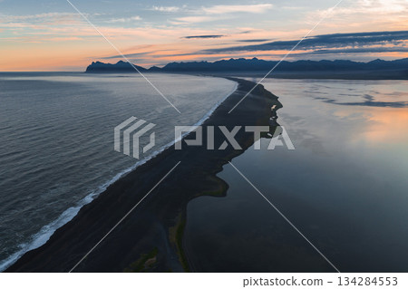 Aerial view of a black sand beach in Iceland, bordered by ocean and calm waters, with rugged mountains and a colorful sunset sky in the background. 134284553