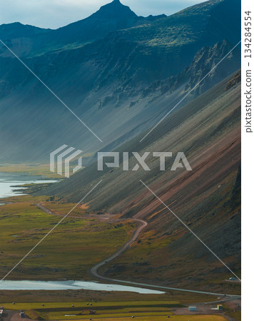 A steep, rugged mountainside in Iceland with earthy tones and green vegetation. A winding road leads to a calm body of water reflecting the peaks. 134284554
