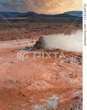 Steaming fumarole surrounded by reddish orange soil in a rocky geothermal area. Rolling hills, snow capped mountains, and a warm sky complete the scene. 134284556
