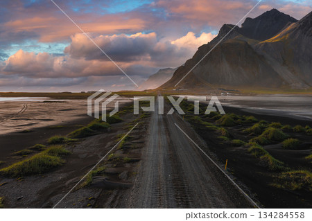 A gravel road winds through black sand toward jagged mountains under a vibrant sunset sky, likely on the Stokknes Peninsula in Iceland. 134284558