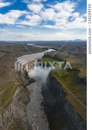 Aerial view of Dettifoss waterfall in Iceland, surrounded by rugged cliffs, a dramatic canyon, rocky terrain, green moss, and a winding river. Aerial view of Dettifoss waterfall in Iceland, surrounded by rugged cliffs, a dramatic canyon, rocky terrain, green moss, and a winding river. 134284559