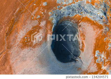 Aerial image of a geothermal area in Iceland with orange and gray tones, featuring a dark oval mud pot, mineral deposits, and steaming earth. 134284571
