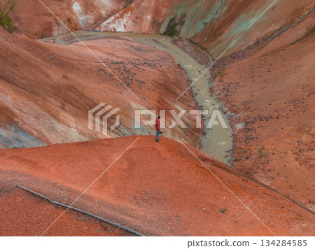 A person in a red jacket stands on a ridge overlooking red and orange toned hills. A winding stream cuts through the rugged, geothermal terrain. 134284585