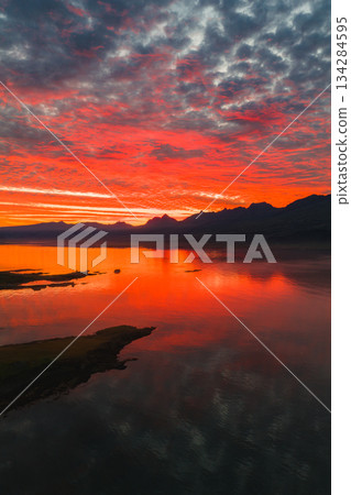 A vivid sunset reflects on calm water in Iceland, with red, orange, and pink hues. Jagged mountains and a grassy peninsula complete the scene. 134284595