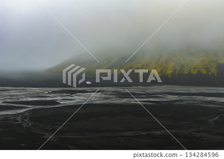 A misty Icelandic landscape with a black sand plain, a moss covered mountain shrouded in clouds, and a distant white vehicle highlighting the vastness. 134284596