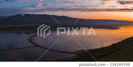Aerial view of Akureyri, Iceland, at sunset with a calm fjord, a curved bridge crossing the water, and snow capped mountains in the background. 134284599