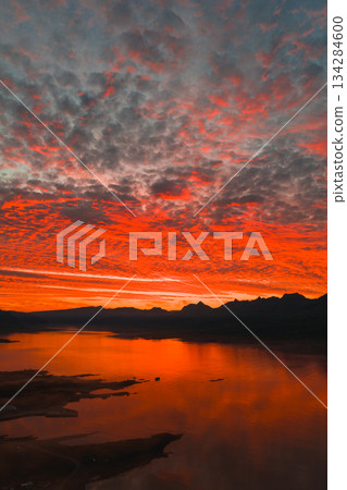 Fiery red and orange sunset reflects on calm water with jagged mountain silhouettes in the background, under a sky filled with textured clouds. 134284600