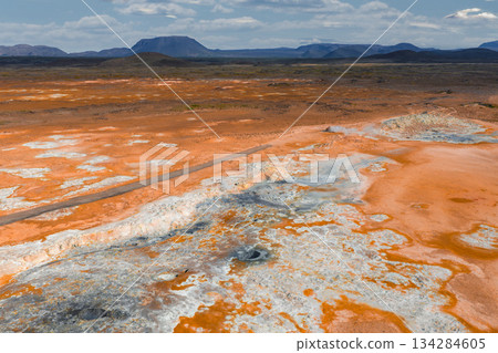 Vibrant geothermal landscape in Iceland with orange and white mineral deposits, steam vents, bubbling mud pots, a road, and rugged mountains under clouds. Vibrant geothermal landscape in Iceland with orange and white mineral deposits, steam vents, bubbling mud pots, a road, and rugged mountains under clouds. 134284605