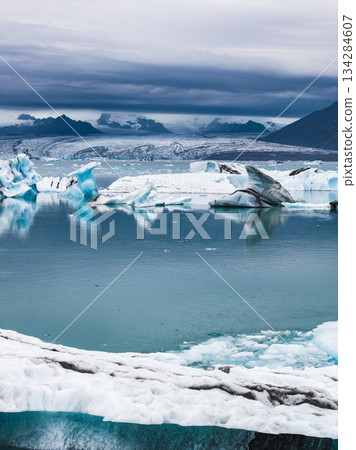 Jokulsarlon Glacier Lagoon features jagged icebergs on calm water, a glacier in the background, rugged mountains, and an overcast sky. Jokulsarlon Glacier Lagoon features jagged icebergs on calm water, a glacier in the background, rugged mountains, and an overcast sky. 134284607