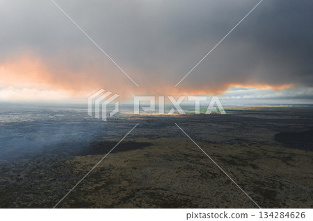 Aerial view of a dark volcanic terrain with mossy patches, illuminated by an orange glow from the sun under thick clouds, with green fields in the distance. Aerial view of a dark volcanic terrain with mossy patches, illuminated by an orange glow from the sun under thick clouds, with green fields in the distance. 134284626