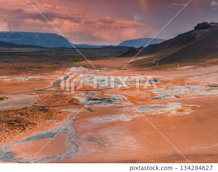 Geothermal area in Iceland with orange and white mineral deposits, steam vents, mud pots, rolling hills, distant mountains, and a winding road. Geothermal area in Iceland with orange and white mineral deposits, steam vents, mud pots, rolling hills, distant mountains, and a winding road. 134284627